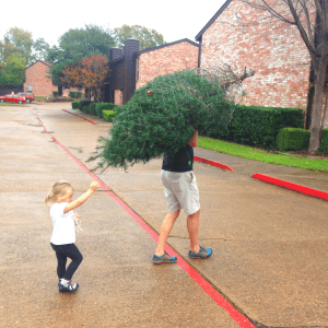 Man carrying real tree on shoulder with little girl trailing behind