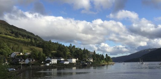 A coastal village in scotland sits on the banks of a loch. There are two sailing boats in the distance.