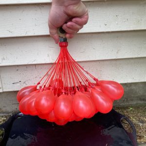 A hand holds an outside tap. Attached to the tap are a set of red water balloons which are almost full of water.