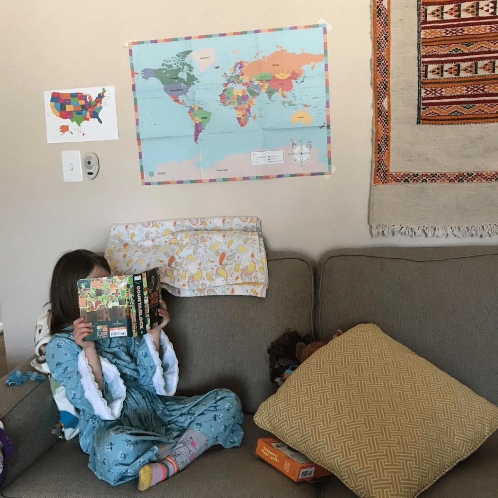 child in blue dress sitting on couch reading a book with maps behind her on the wall