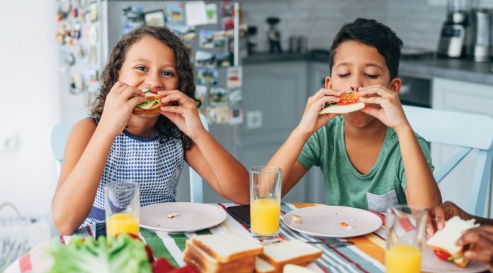 two children sitting at a table and eating sandwiches