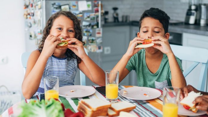 two children sitting at a table and eating sandwiches