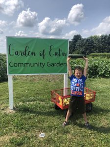 boy in blue shirt stands in front of green sign saying Garden of Eat'n while holding a zucchini above his head