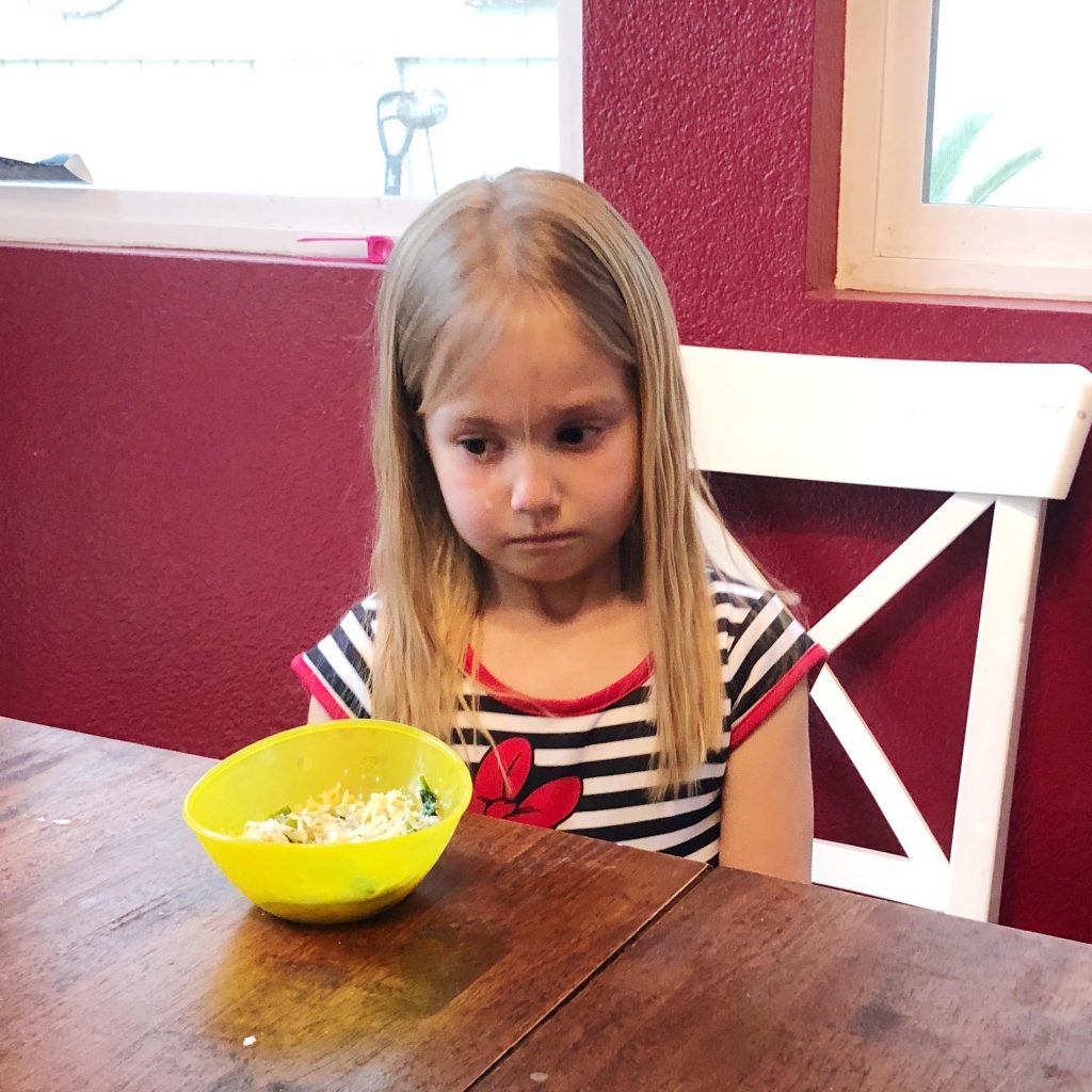 small girl, tearfully sitting at a table with a yellow bowl of food in front of her