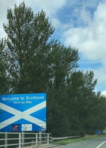 Large roadside sign says 'Welcome to Scotland'. There are large trees behind the sign.