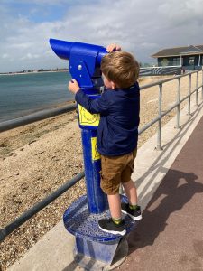 A boy stands looking into a telescope on the side of the beach, which is pointing out to sea.
