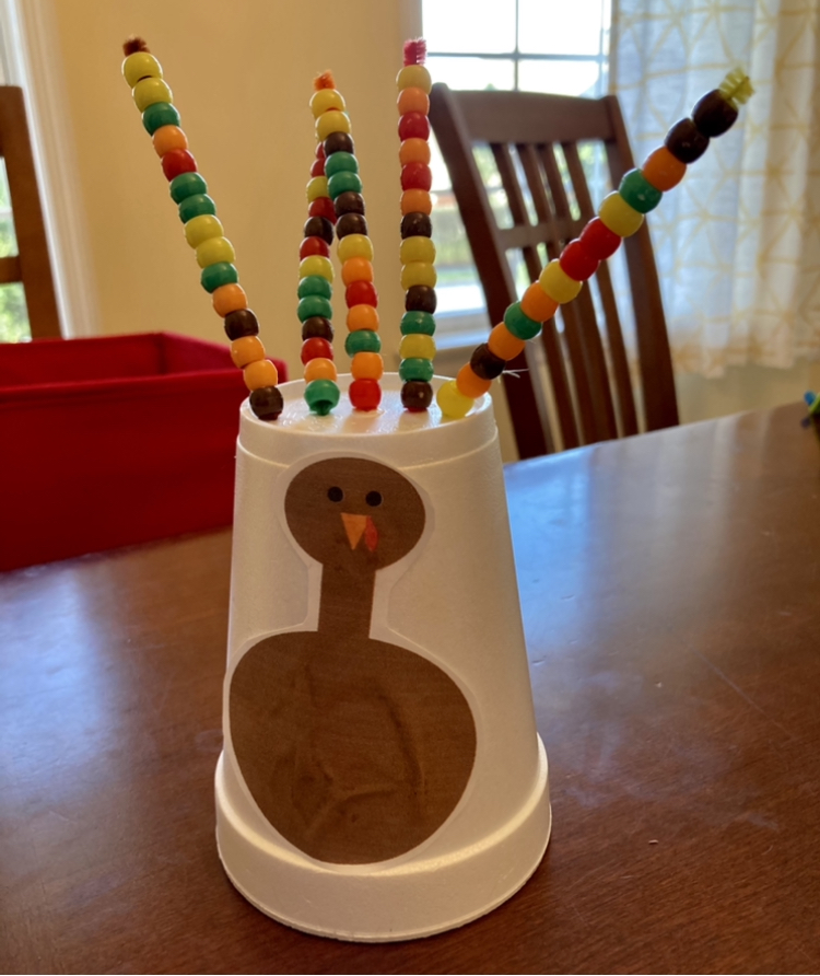 A styrofoam cup is upside down on the table with a picture of a turkey on it. The feathers are made out of pipecleaners sticking out of the cup with multi-coloured beads threaded onto them.