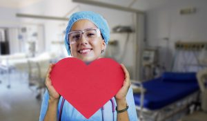 nurse in scrubs holding up a red heart in a hospital room