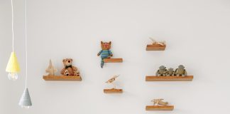 Natural wood Montessori-style toddler bed with pillows, small blanket. Toys sit on shelves mounted high above the bed.