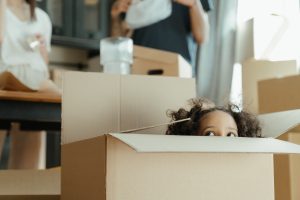 child peeking out of a moving box while parents are in the back ground unpacking