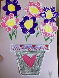Handmade Mother's day card. Lilac background with purple and pink flowers drawn and stuck on the card. They are in a vase with a heart on. Card says 'Mummy and Me'.