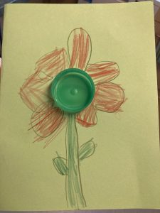 Mother's day card. Green plastic bottle lid stuck to yellow card. Lid forms middle of the flower which has been drawn around it. The flower has red petals and green stem.