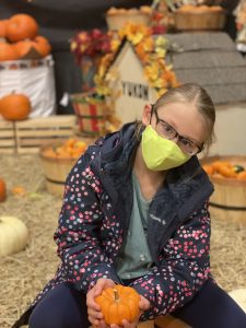 A girl wearing a face mask holds a small pumpkin. There is a pumpkin patch and fall decorations in the background. 