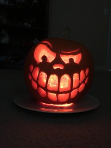 A carved pumpkin is in the dark. A light shines through highlighting large eyes and a wide, sinister smile.