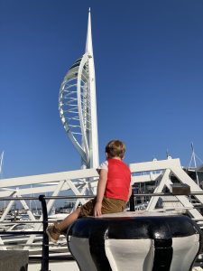 A young boy (4) sits in the foreground looking away from the camera. In the distance, the white Spinnaker Tower stands out again a bright blue sky.