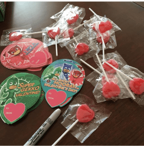 PJ Mask valentine cards lie on a table with hear-shaped lollies beside them. A sharpie is also in view.