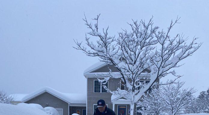 Man using snow blower to clear walk way in several feet of snow.