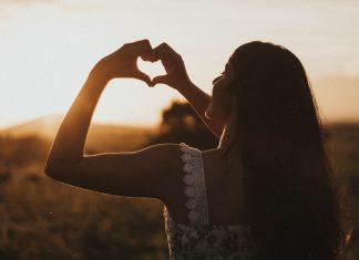 The silhouette of a woman can be seen against the setting sun. She is holding up her hands, forming a heart shape with her fingers.