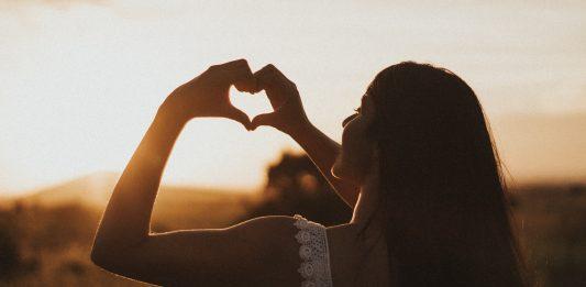 The silhouette of a woman can be seen against the setting sun. She is holding up her hands, forming a heart shape with her fingers.