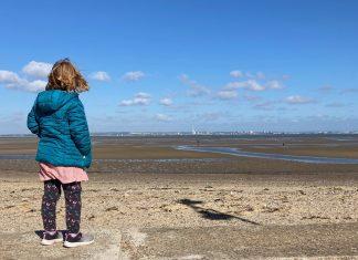 A young girl stands in the foreground, facing away from camera. She is looking out over a deserted beach on a clear day.