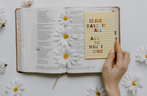A hand rests on the bible holding a paper with different colored letters. The paper reads "Jesus paid it all. All to Him I owe." White daisies with yellow centers surround the bible.