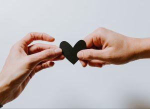 Two hands are holding a black paper heart, each holding one side.