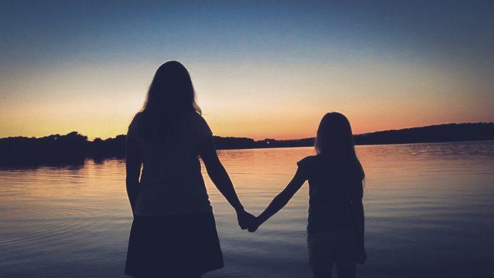 A silouette of a woman and young girl are seen, facing away from camera towards a sunset in the distance. They are holding hands.