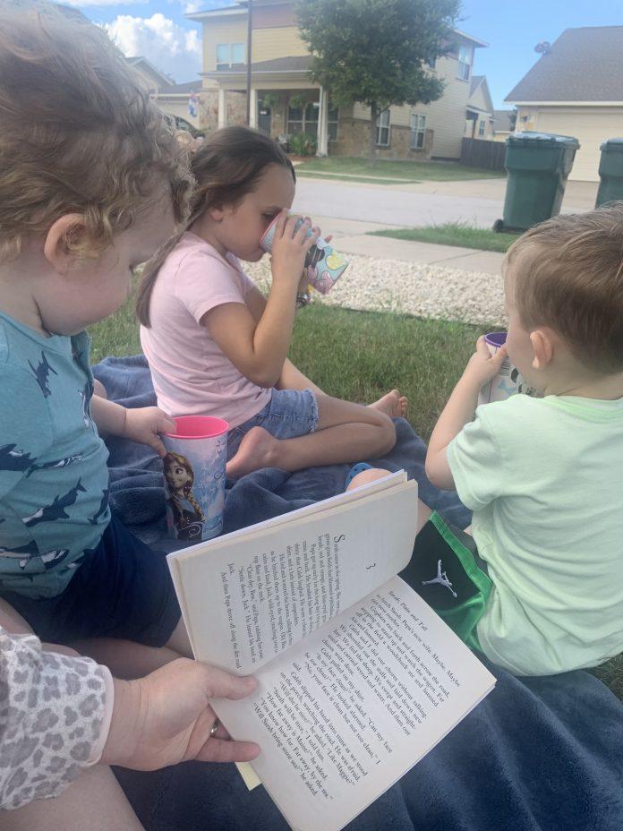 Photo of three young children sitting outside on a blue blanket, drinking out of plastic cups. The book, "Sarah, Plain and Tall" is opened next to them and is being read aloud by the mother, who is not in the photo.