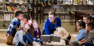 An adult woman wearing a blue sweater in a library reading to a small group of diverse children and a service animal