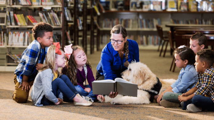 An adult woman wearing a blue sweater in a library reading to a small group of diverse children and a service animal